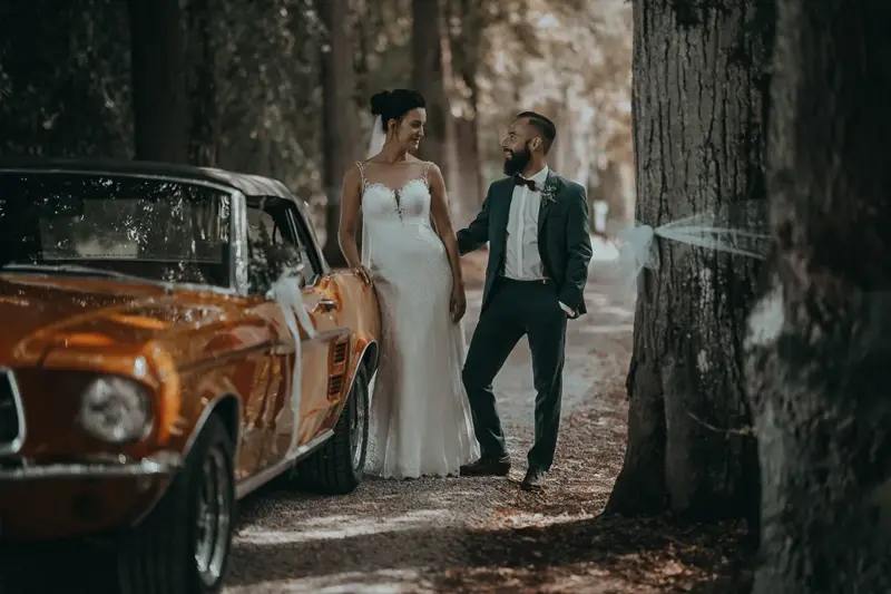 Ein Hochzeitspaar neben einem orangefarbenen Oldtimer im Wald